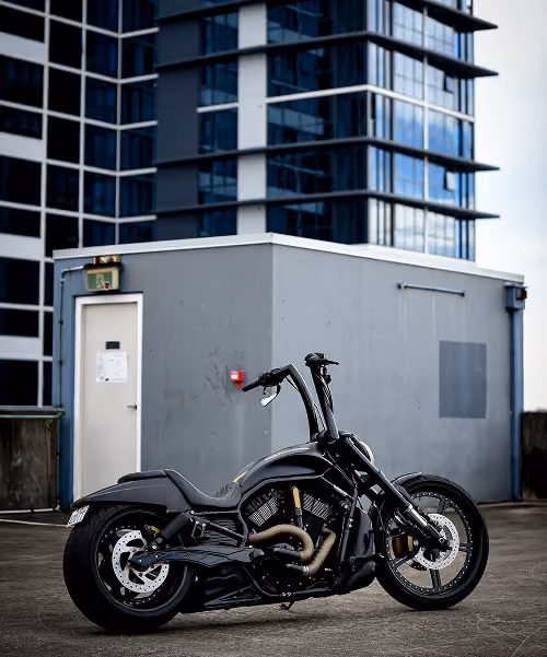 Black cruiser motorcycle parked on concrete near a gray utility building with a modern glass skyscraper background.