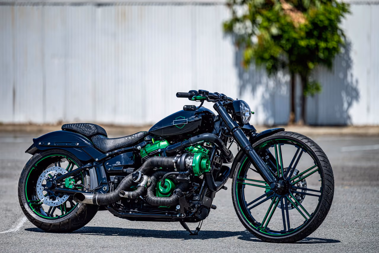 Black custom motorcycle with green engine and wheel accents parked on pavement near a white building.