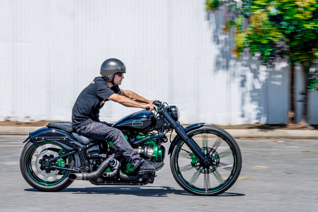 Man wearing helmet riding a black and green custom motorcycle on a street beside a white fence.