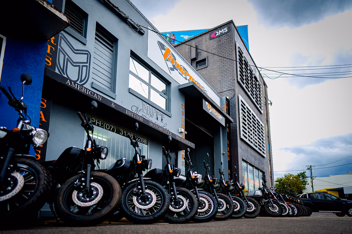 Line of black motorcycles parked outside a gray building with motorcycle shop signs.