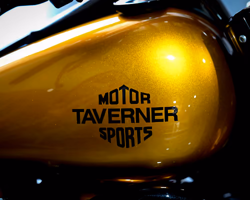 Close-up of a golden motorcycle fuel tank with black text 'Motor Taverner Sports' on it.
