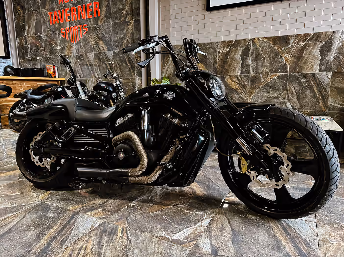 Black custom motorcycle parked indoors on tiled floor with tavern sports sign in background.