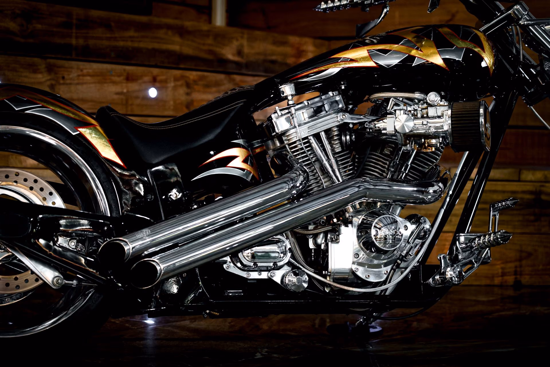 Black custom motorcycle with chrome engine parts and dual exhaust pipes against wooden background.
