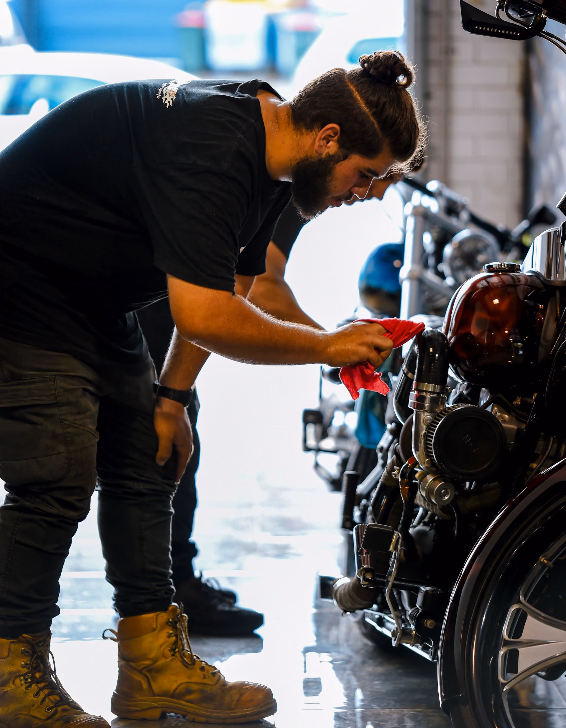 Bearded man in black shirt cleaning a motorcycle engine with a red cloth inside a garage.