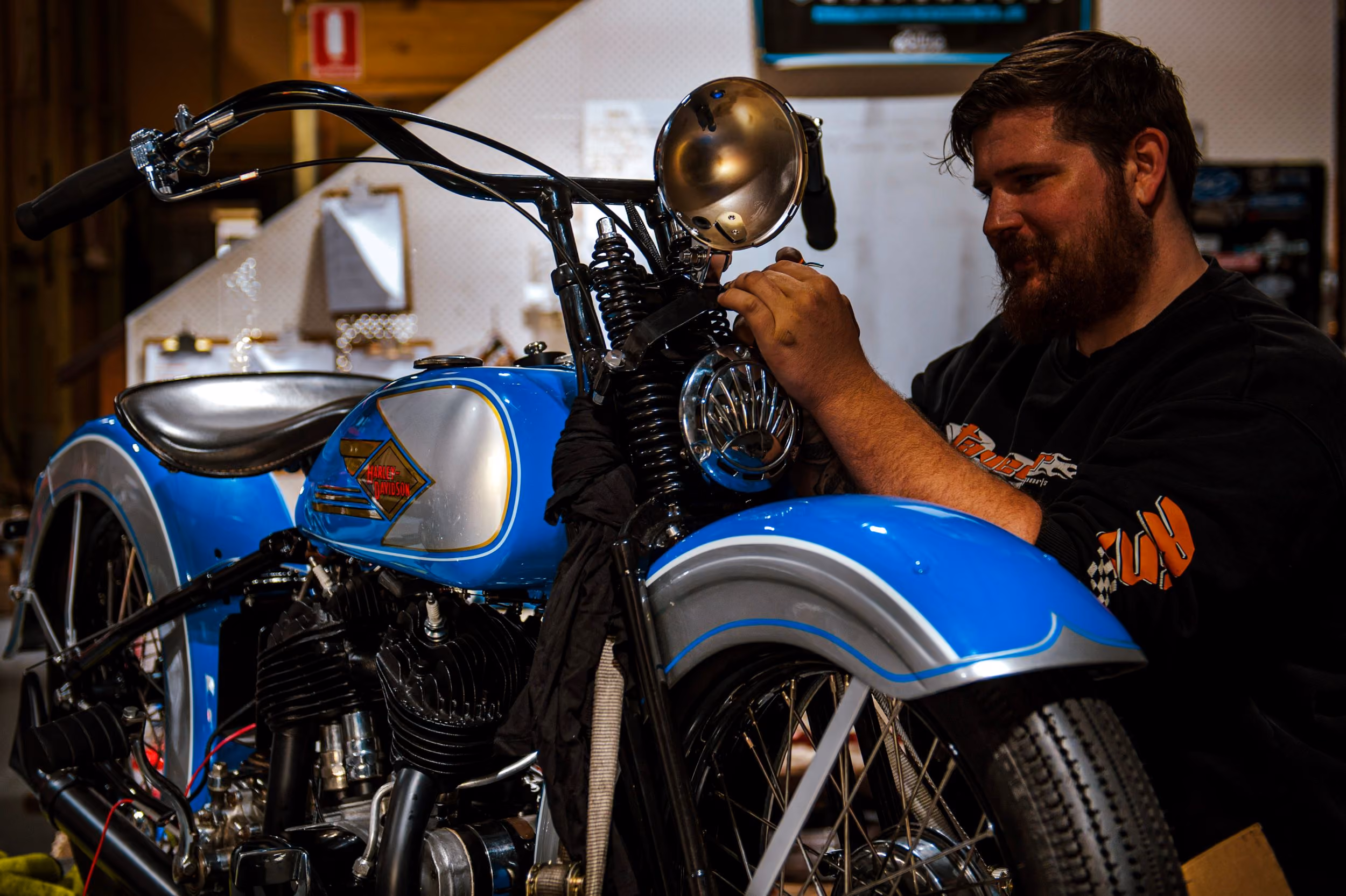 Bearded man in black shirt fixing the headlight of a vintage blue Harley-Davidson motorcycle indoors.