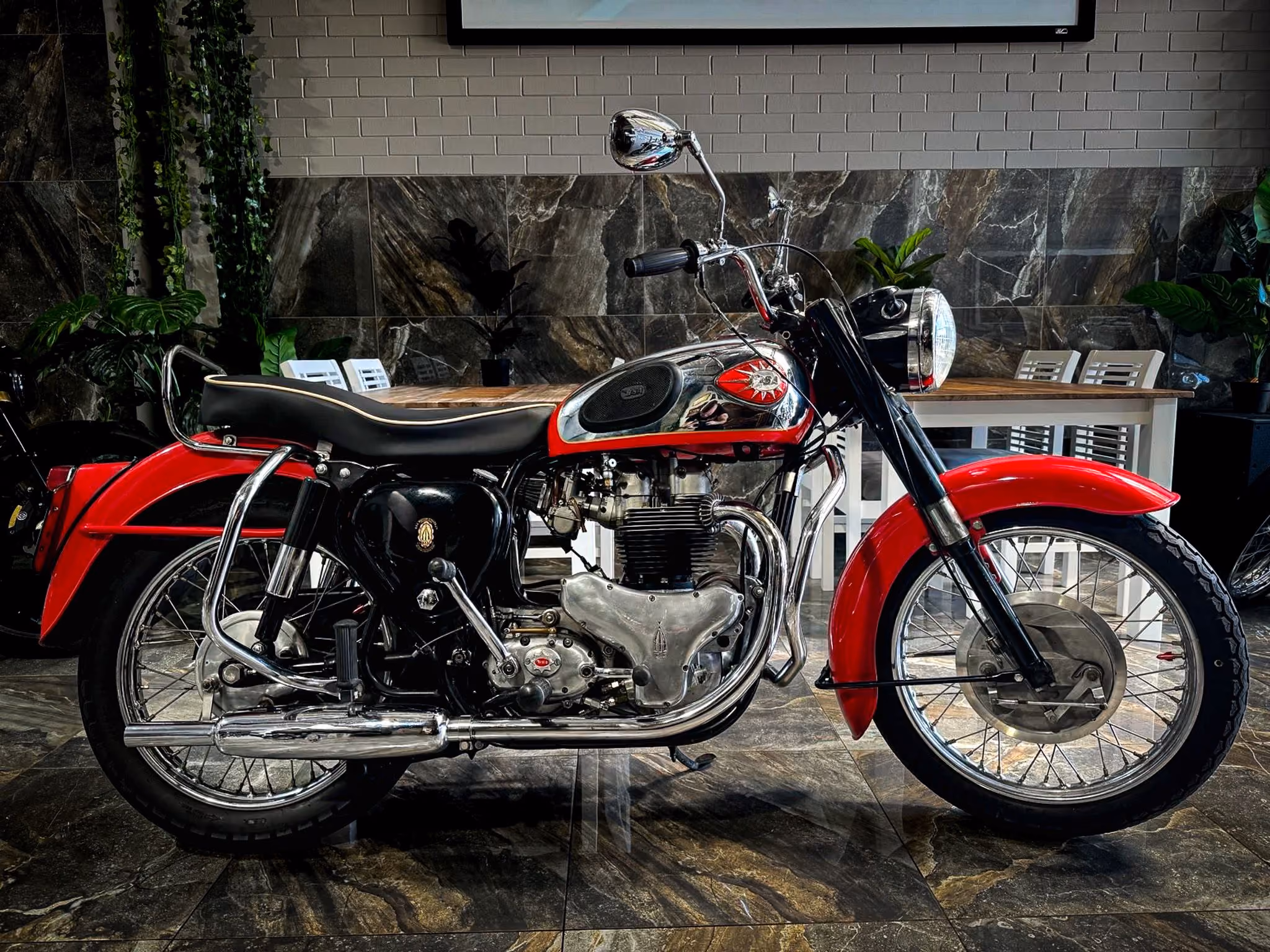 Classic red and black vintage motorcycle parked indoors on marble floor with plants and table in background.