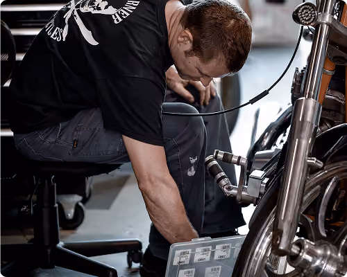 Man working on a motorcycle, seated on a wheeled stool, inspecting parts closely.