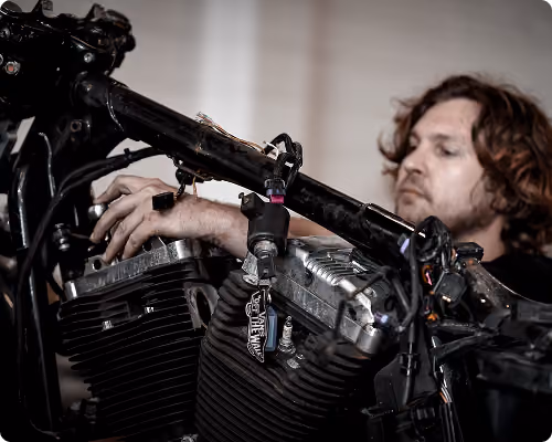 Man with curly hair working on a motorcycle engine frame, focusing on engine parts and wires.