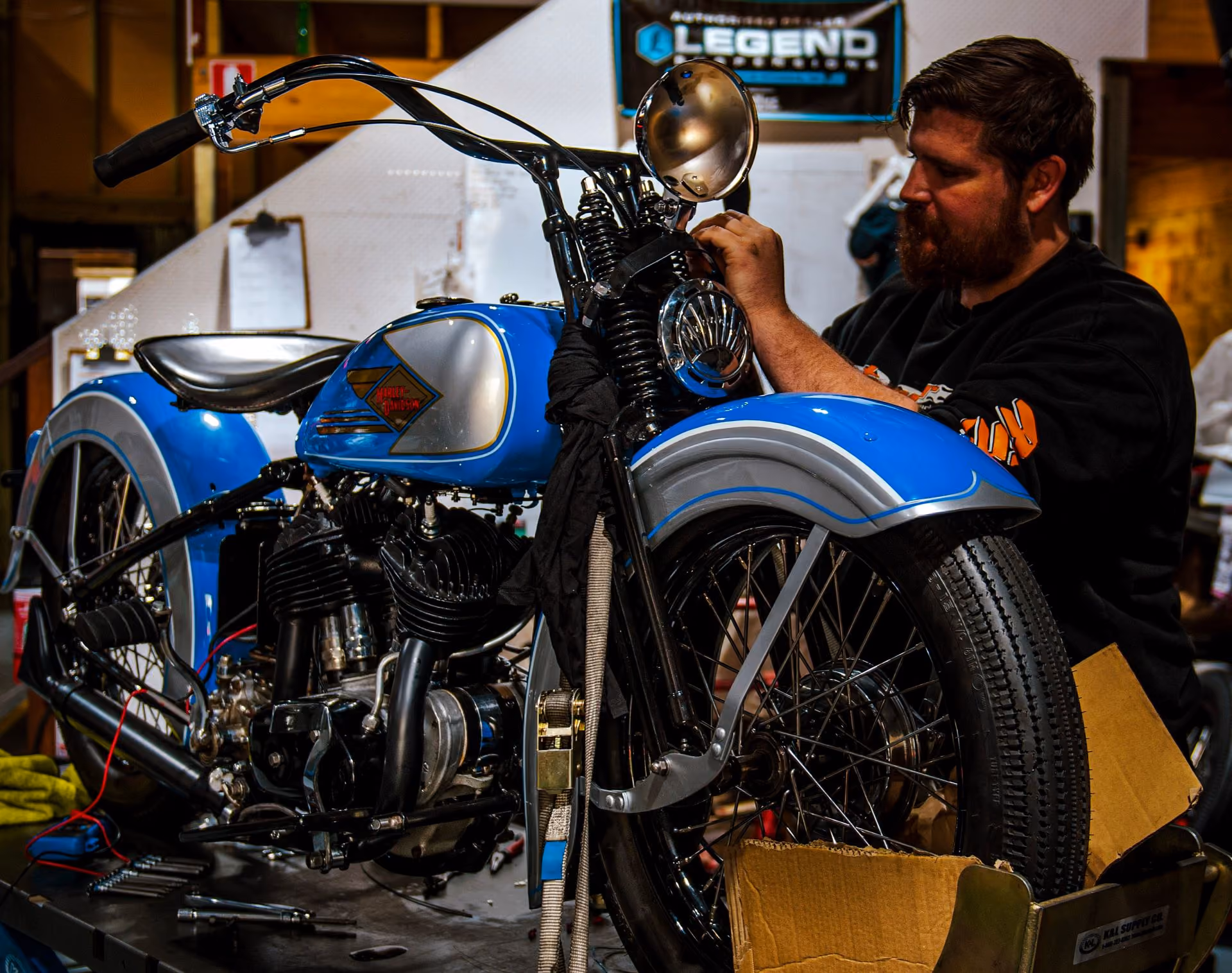 Man with beard working on a vintage blue Harley-Davidson motorcycle in a workshop.