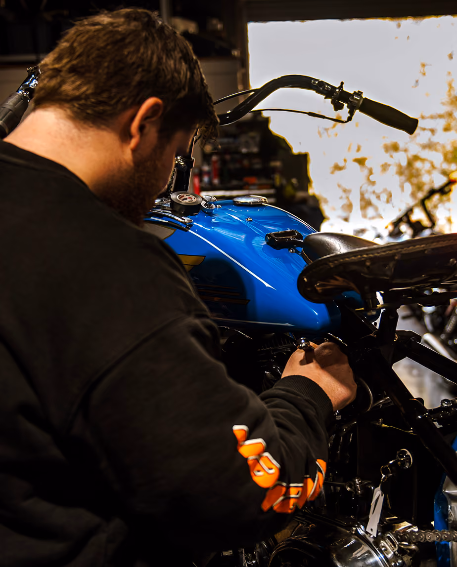 Man in black sweatshirt working on a blue motorcycle in a dimly lit garage.