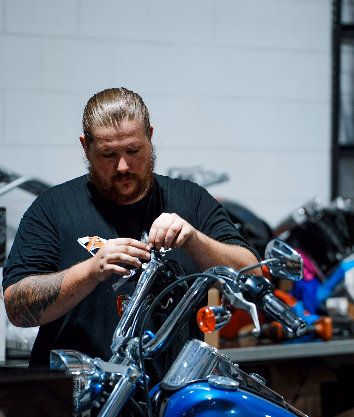 Man with a beard working on the handlebars of a blue motorcycle in a workshop.