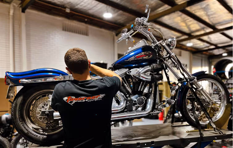Man working on a blue custom motorcycle inside a garage with a metal roof and white brick wall.