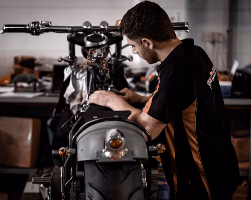 Man in black and orange shirt working on the wiring of a motorcycle in a workshop.