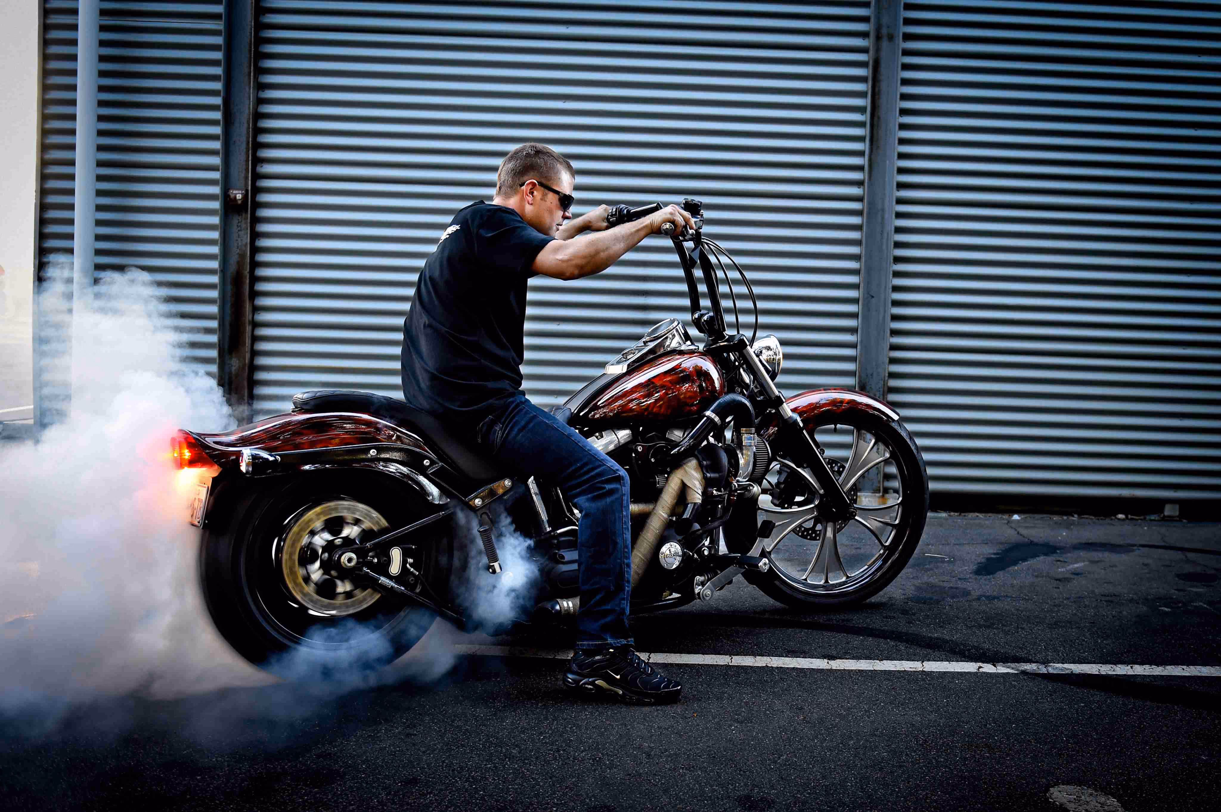 Man wearing sunglasses performs a burnout on a custom motorcycle, creating smoke behind the rear wheel.