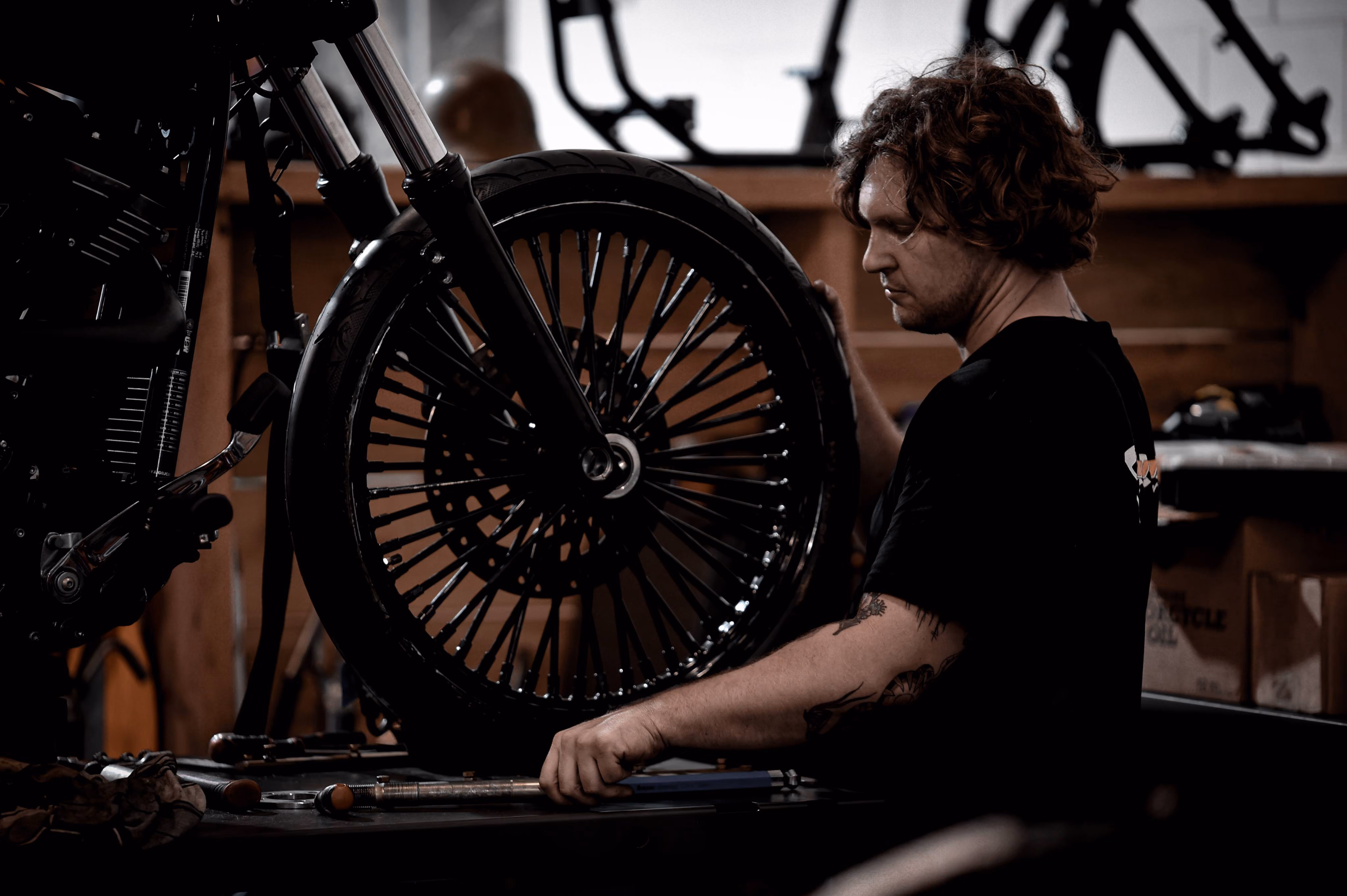 Man with curly hair repairing a motorcycle wheel in a workshop with tools on the table.