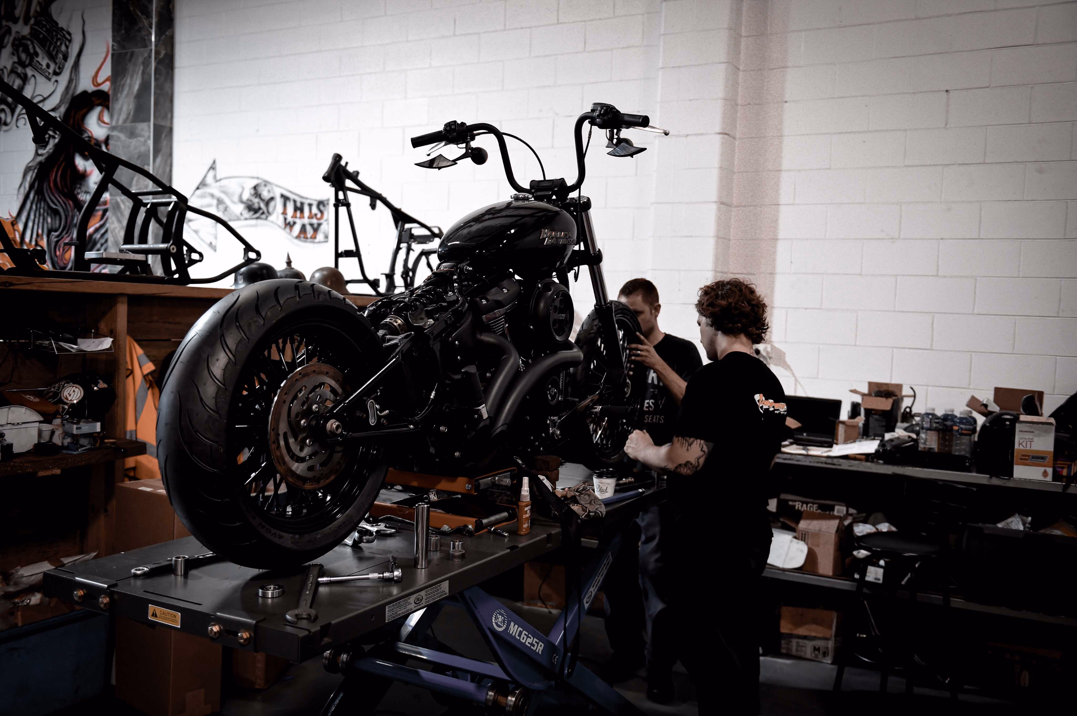 Two men working on a black motorcycle in a workshop with tools and motorcycle parts around.