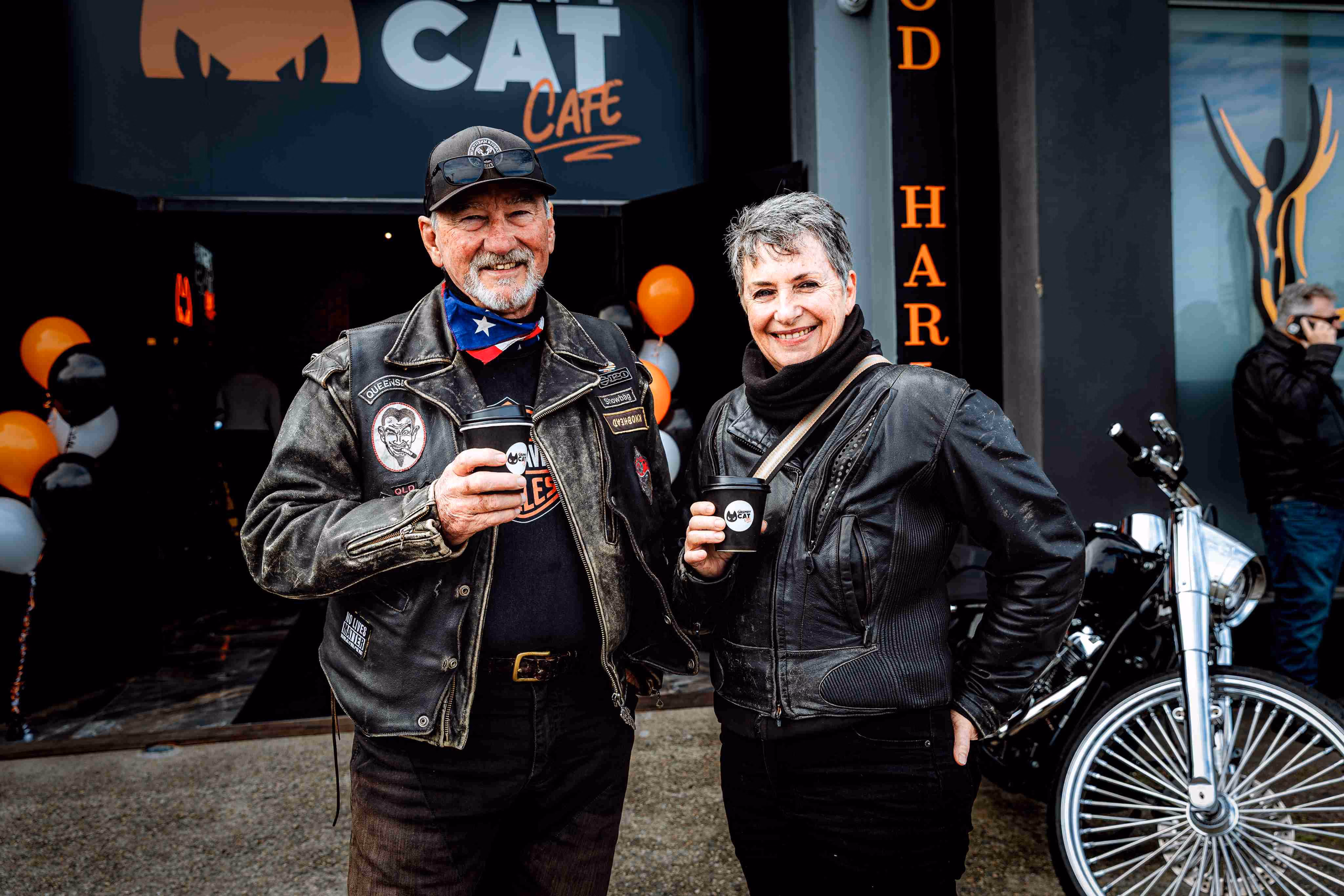 Two smiling people in leather jackets holding coffee cups outside The Grumpy Cat Cafe with a motorcycle nearby.