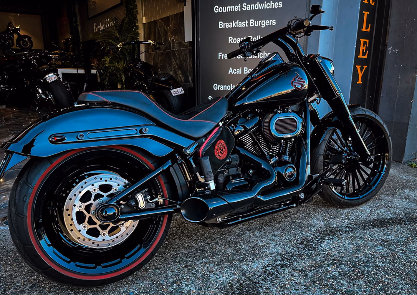 Custom black motorcycle with red accents parked on concrete near a gourmet sandwich menu sign.