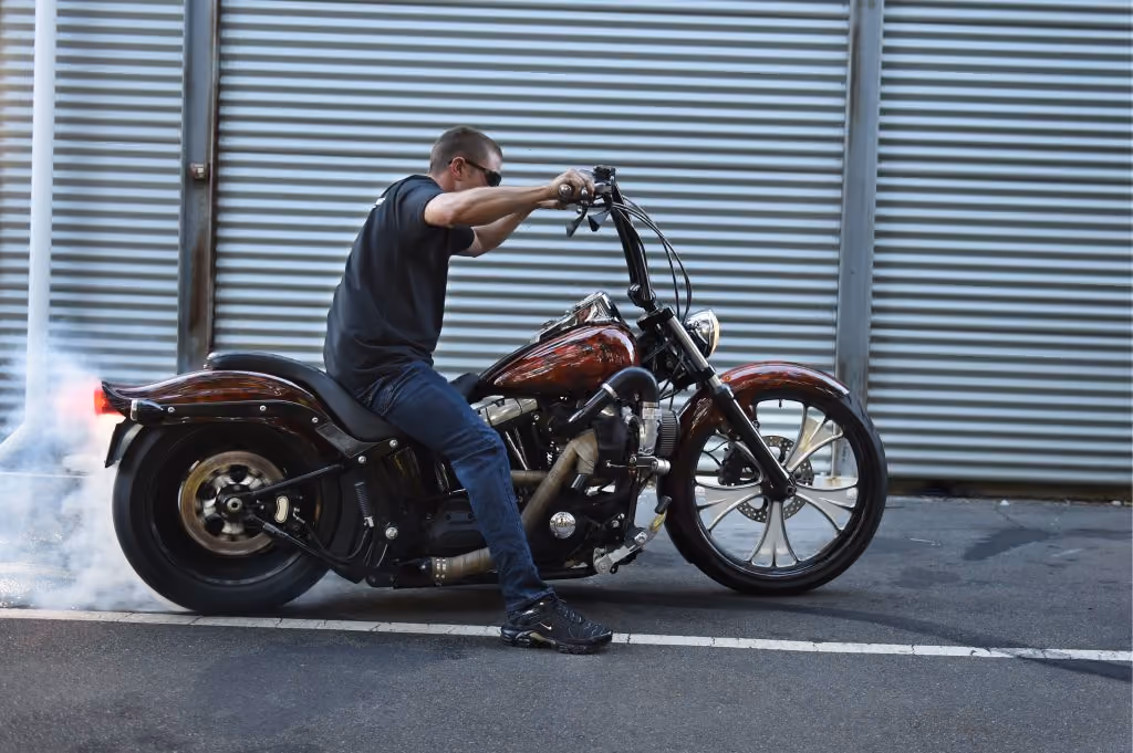 Man in black shirt and sunglasses doing a burnout on a red custom motorcycle on asphalt.