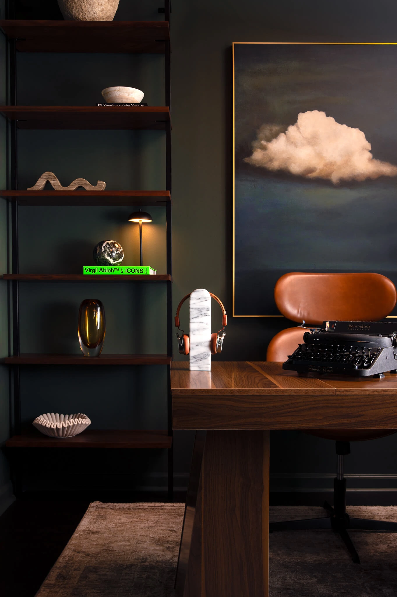 Stylish office desk with a marble headphone stand, a vintage typewriter, a brown leather chair, and a large painting of a white cloud on a dark wall.