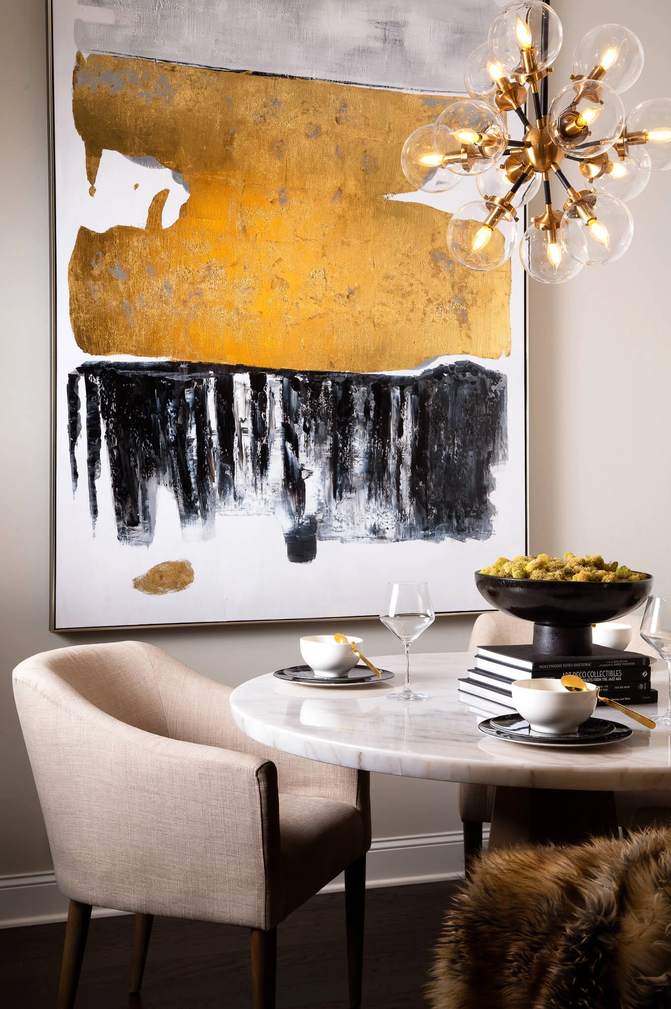 Modern dining room with a round white marble table, beige upholstered chairs, abstract gold and black wall art, and a chandelier with exposed bulbs.
