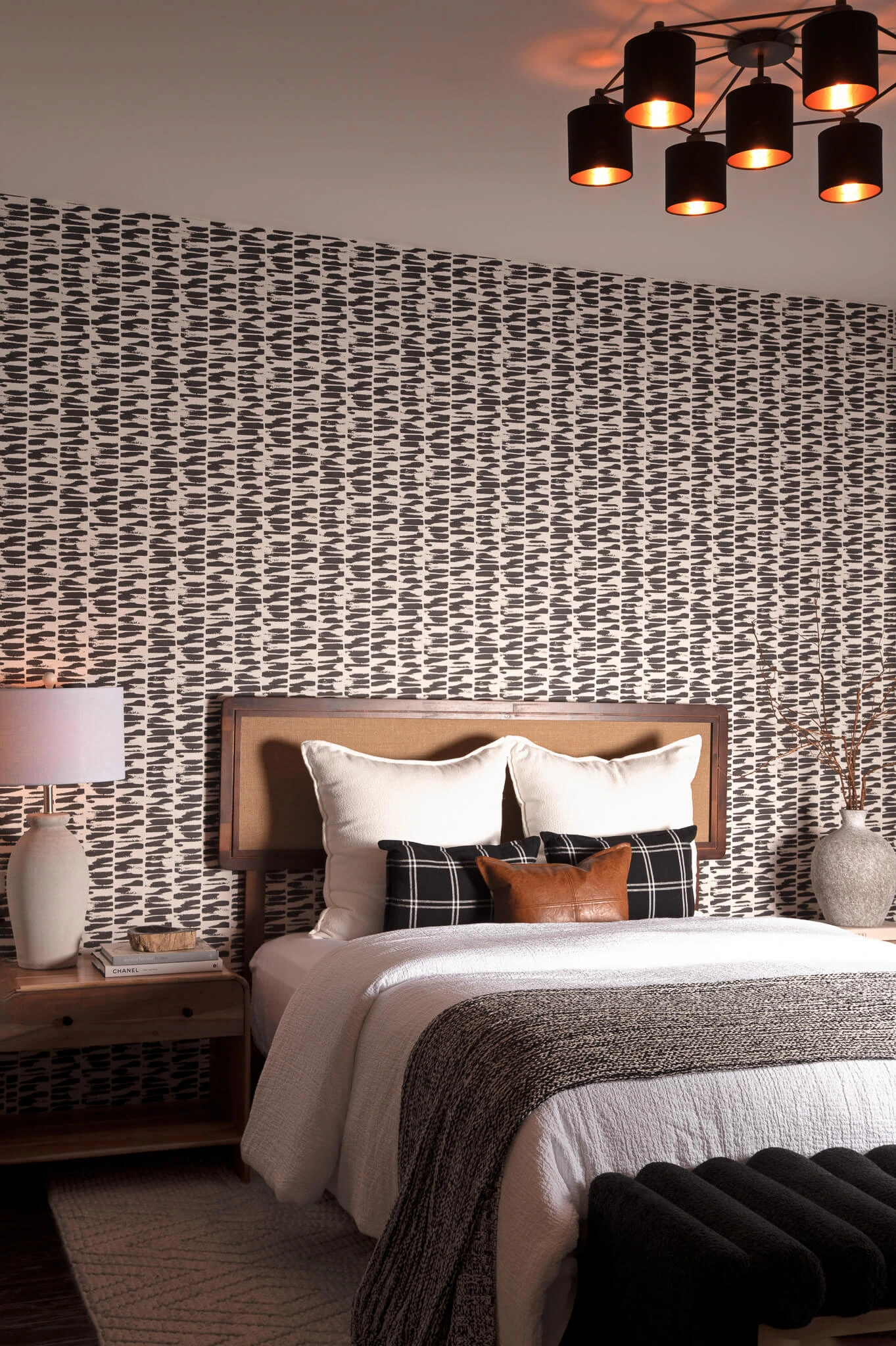 Cozy bedroom with a wooden headboard, white bedding, patterned pillows, a black bench, and modern pendant lighting.