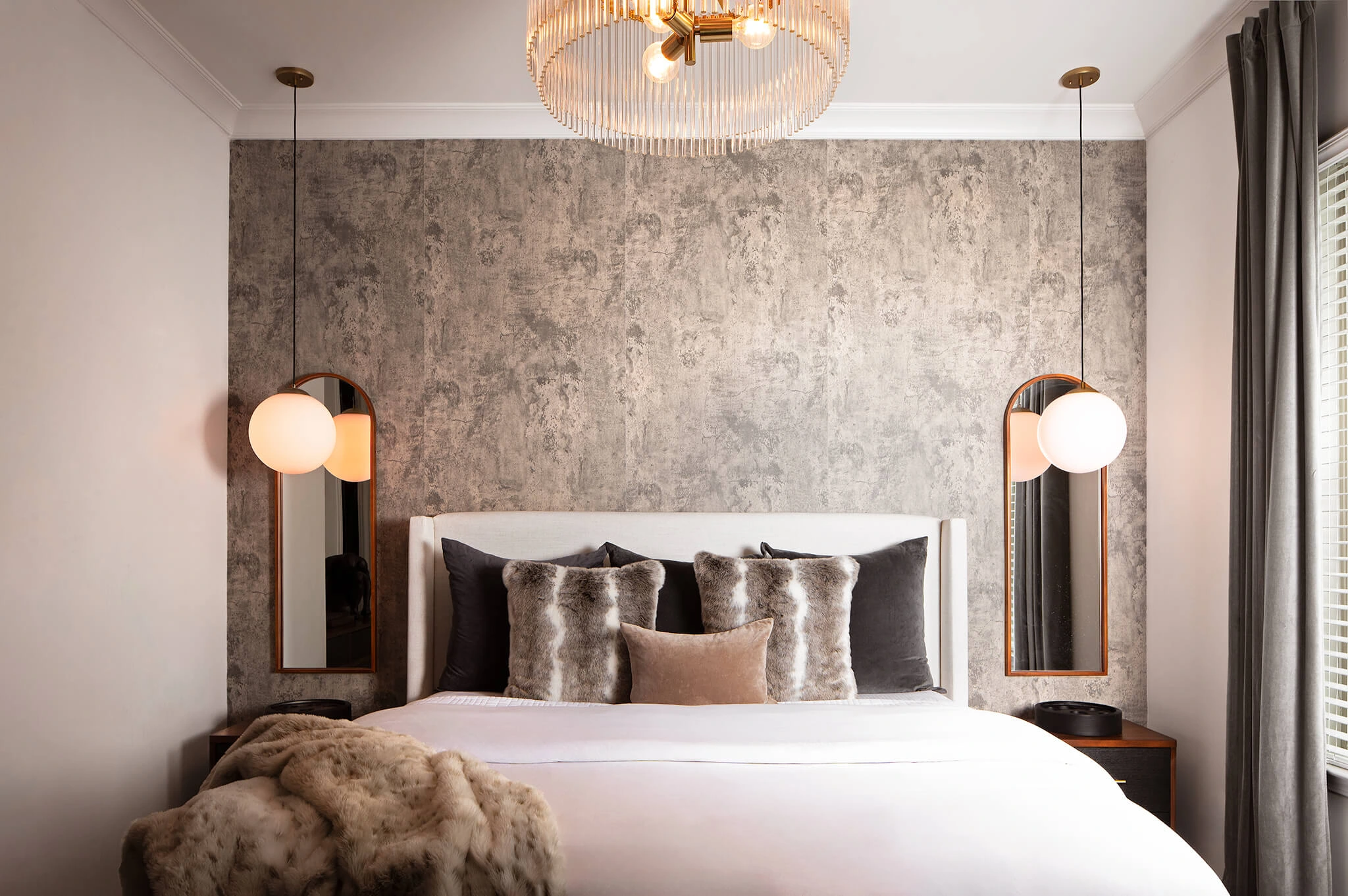 Modern bedroom with a white upholstered bed, gray textured accent wall, fur and velvet pillows, hanging globe pendant lights, and a plush fur throw.