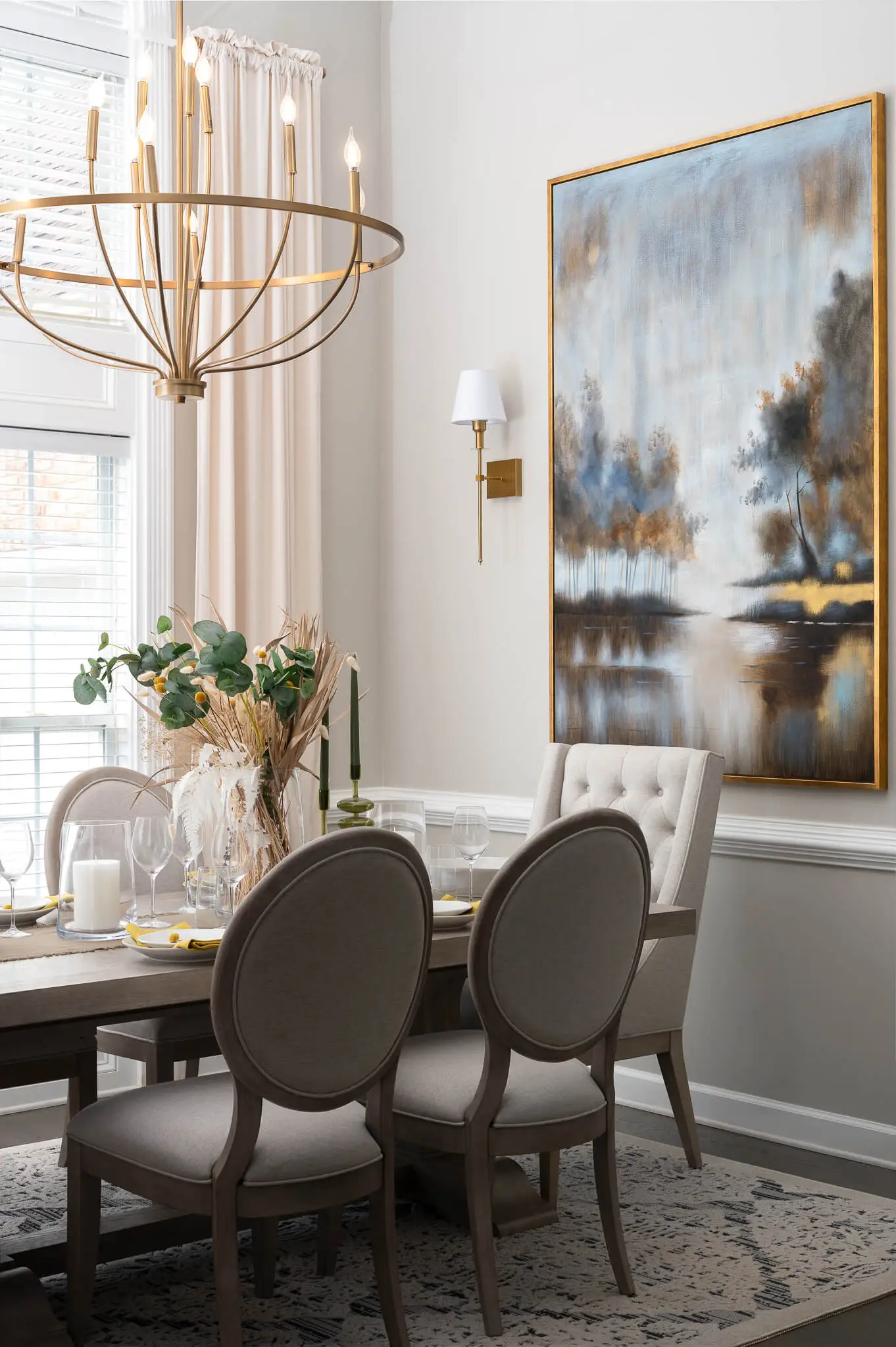 Elegant dining room with a wooden table set with glassware, cream upholstered chairs, a large landscape painting, and a gold chandelier.