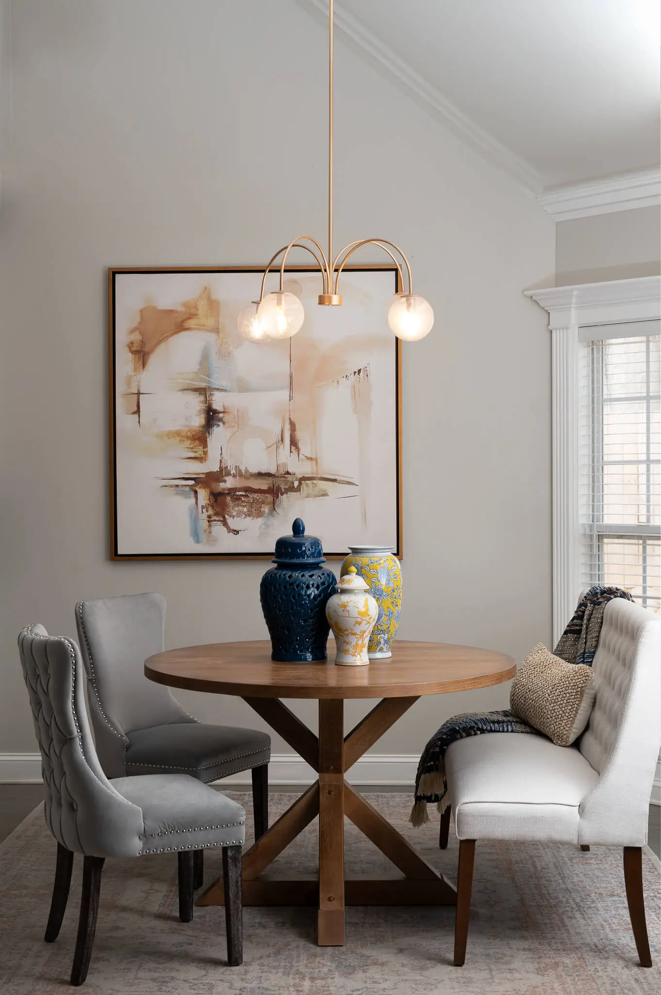 Round wooden dining table with three decorative vases, surrounded by two gray upholstered chairs and a white bench with pillows and a throw, under a modern chandelier in a softly lit room.