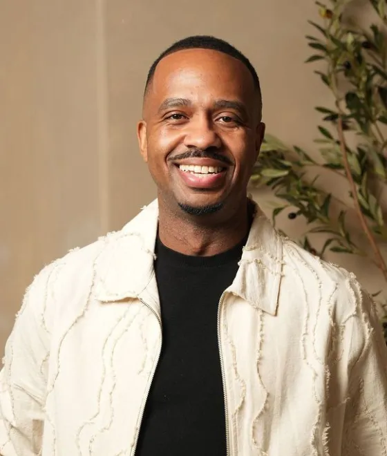 Smiling man wearing a white textured jacket over a black shirt against a neutral background with a leafy plant on the right.