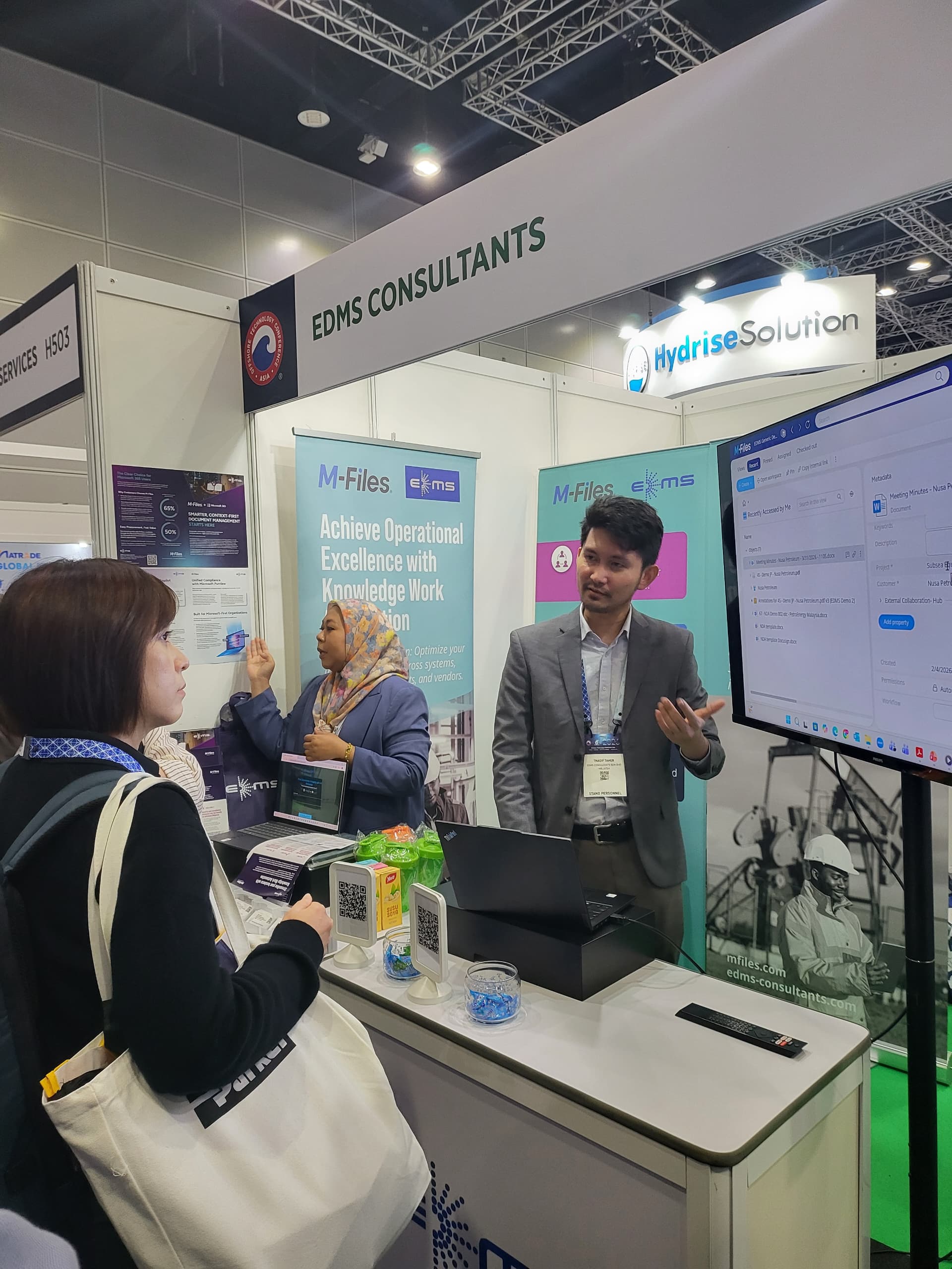 Three people interact in an indoor event space; a man holds a Lenovo laptop while a woman wearing a headscarf smiles, and another woman with a headscarf talks on a phone holding bags.