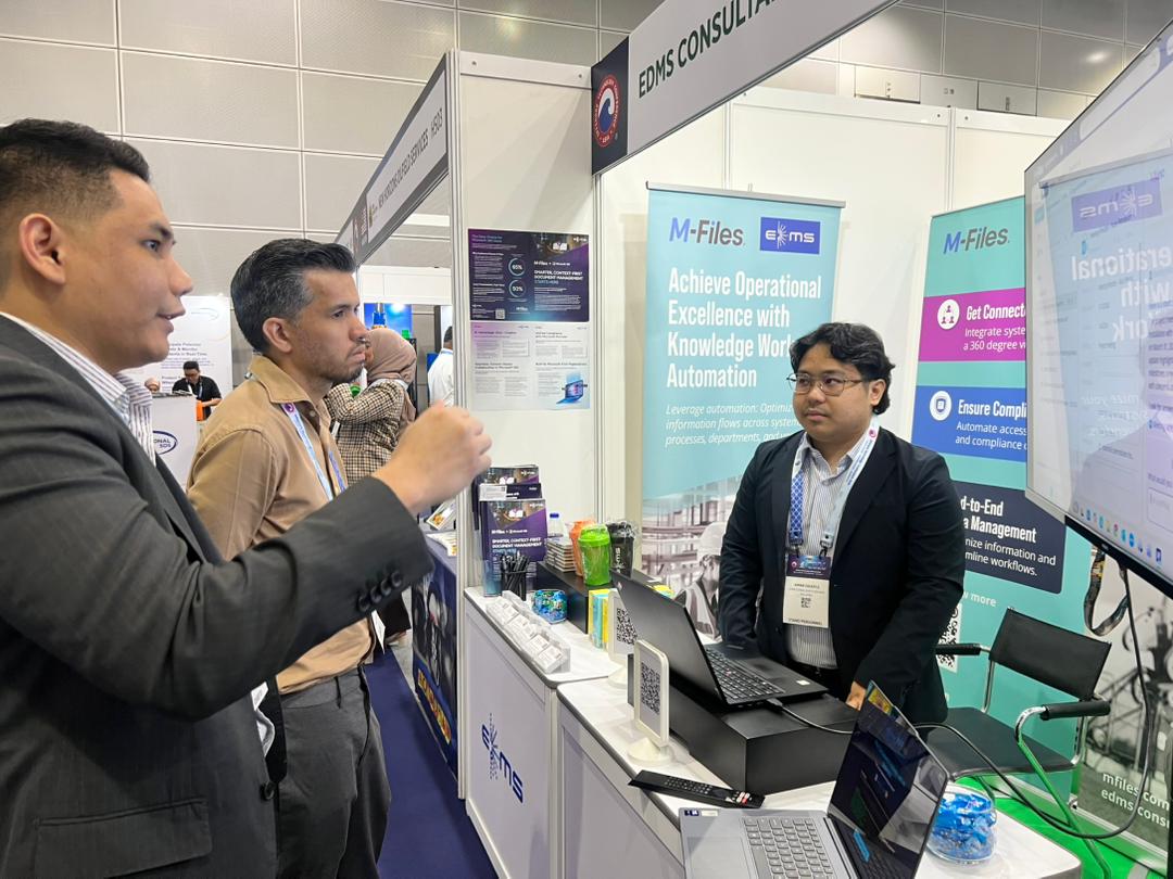 Two men in business attire smiling at a booth table with a laptop and black gift bags, while a man and woman talk in the background at a conference.
