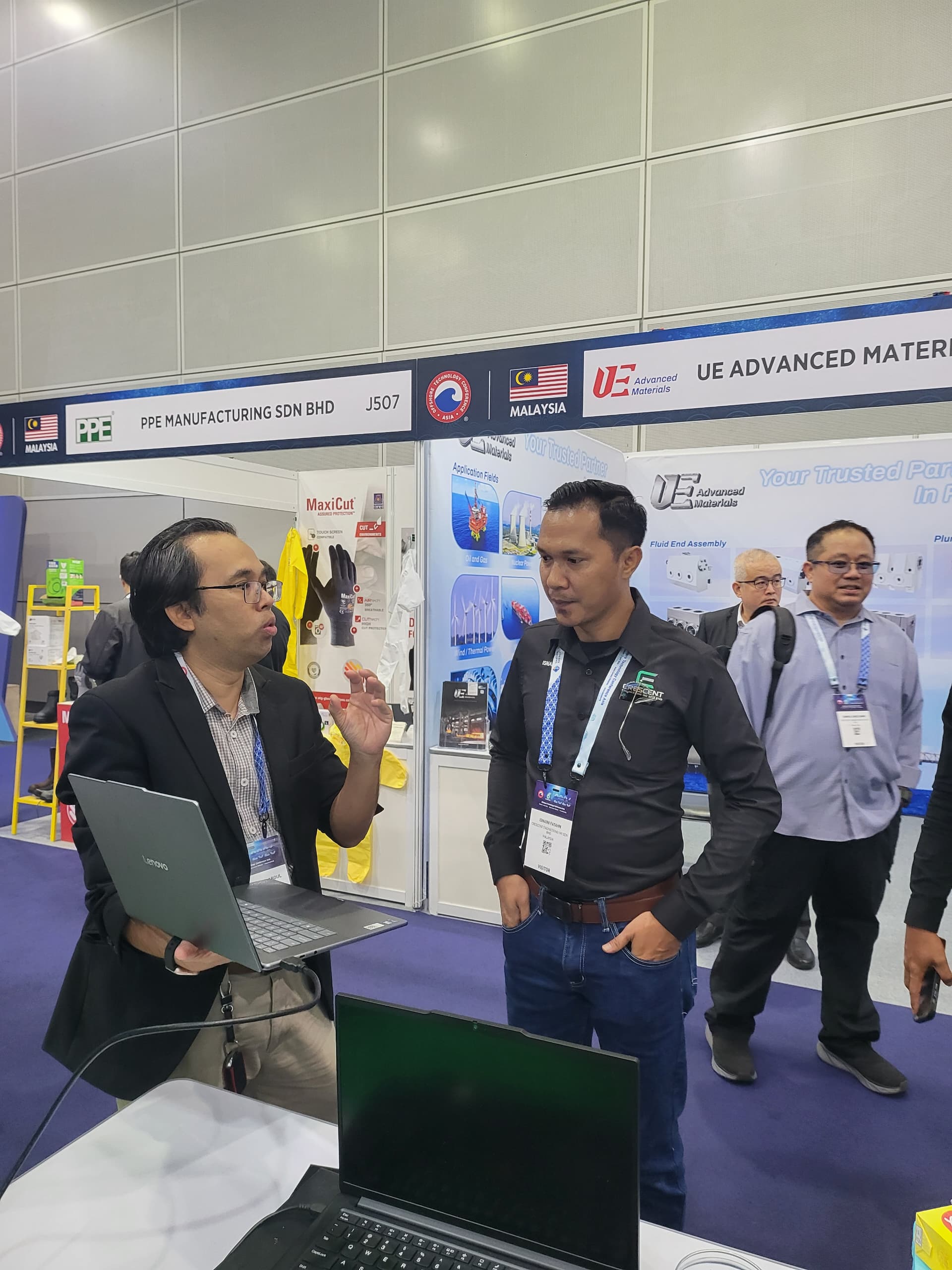 A group of eight people smiling and giving thumbs up behind a table with a laptop and promotional materials at an event.