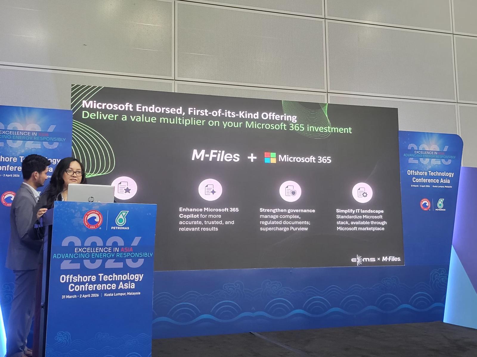 Man in a dark suit and glasses standing and holding a brochure at an exhibition booth with M-Files and EKMS branding.