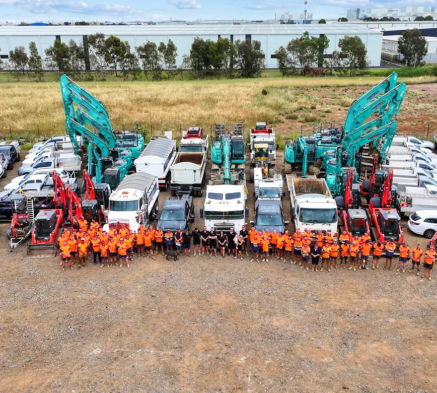 Large group of workers in orange and black shirts standing in front of a fleet of various construction vehicles, including excavators, dump trucks, and vans, parked on a dirt lot with industrial buildings and trees in the background.