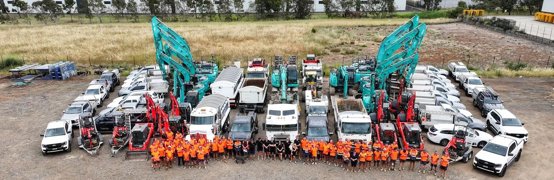 Group of construction workers in orange shirts standing in front of various construction vehicles and machinery in an outdoor lot.