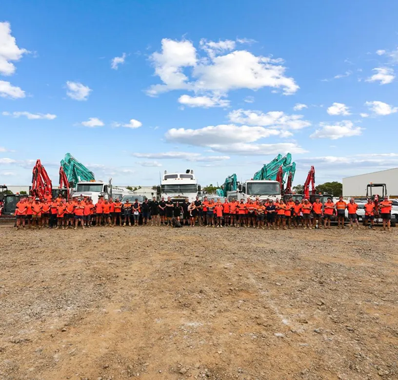 Large group of construction workers in orange and black uniforms standing in front of trucks and excavators on a dirt lot under a blue sky with clouds.