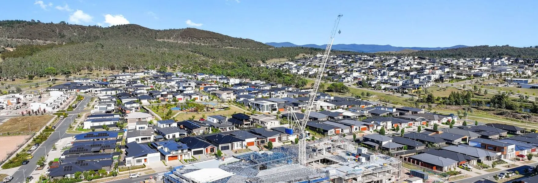 Aerial view of a suburban neighborhood with modern houses, a construction site with a crane, and surrounding green hills under a clear blue sky.