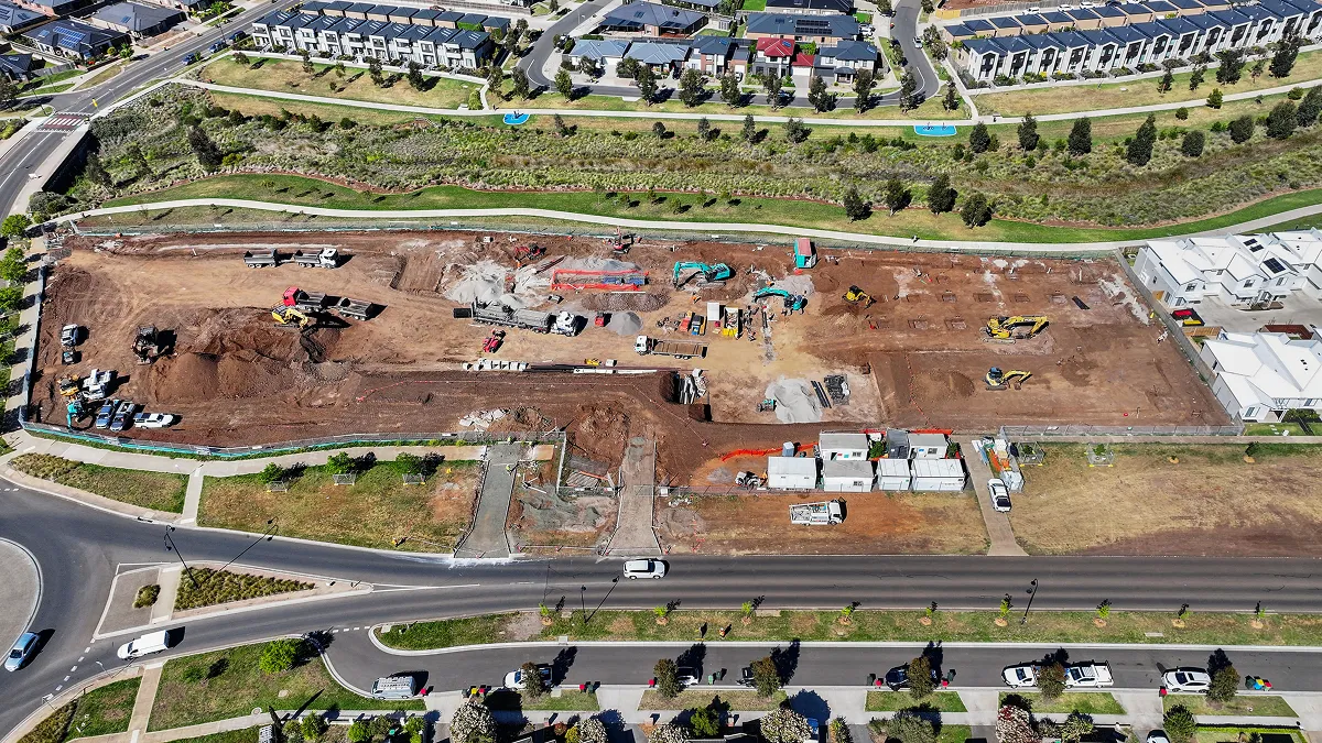 Aerial view of a construction site with earthmoving equipment and trucks, surrounded by residential houses and roads.