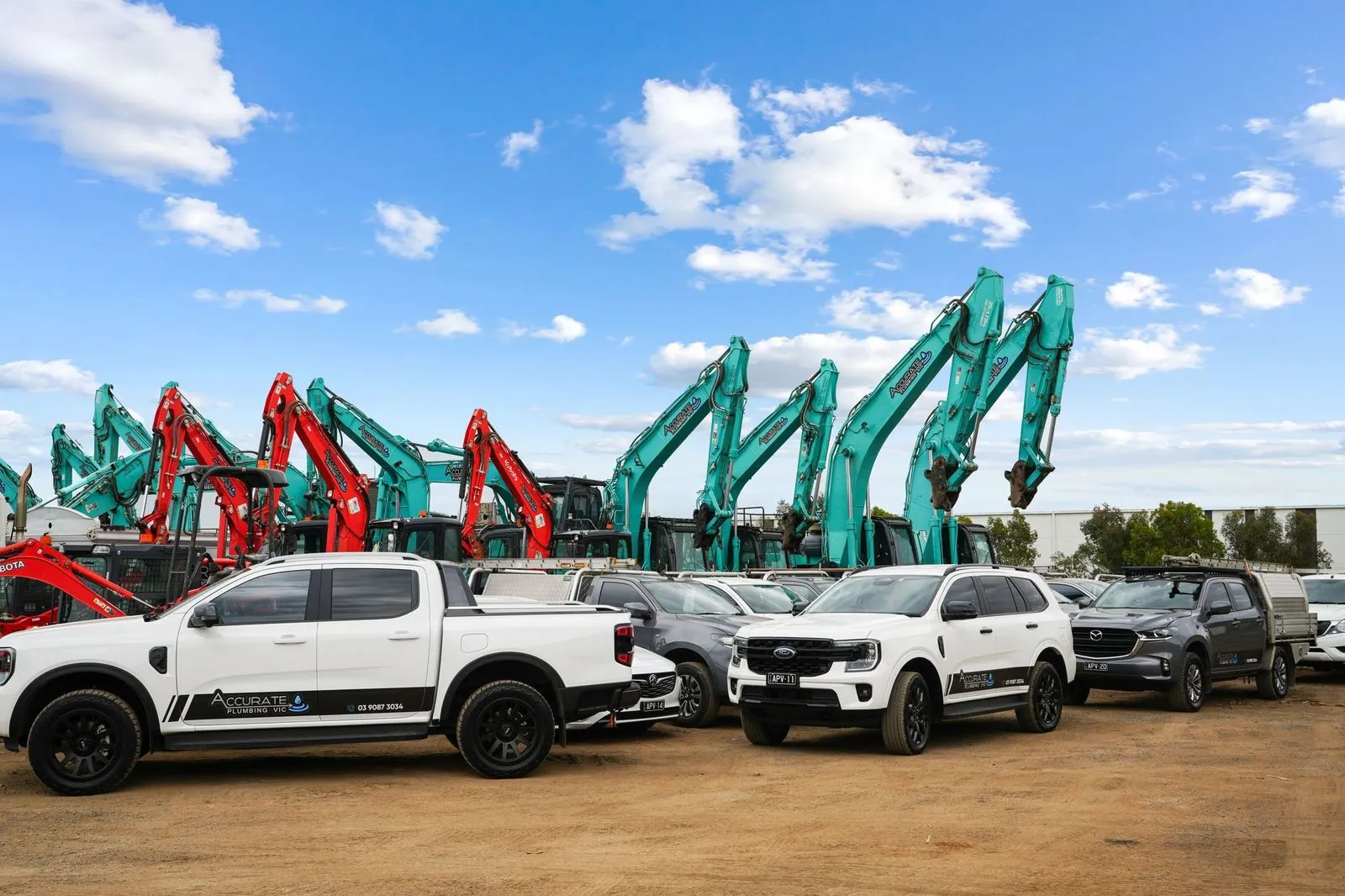 Row of white and gray pickup trucks and SUVs with Accurate Plumbing VIC branding parked in front of green and red excavators under a partly cloudy blue sky.