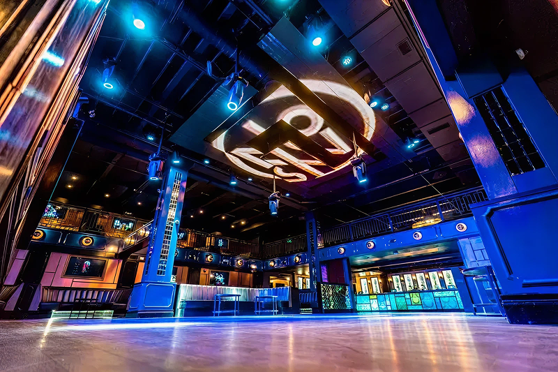 Wide-angle view of an empty nightclub interior with blue lighting and a large illuminated ceiling logo.