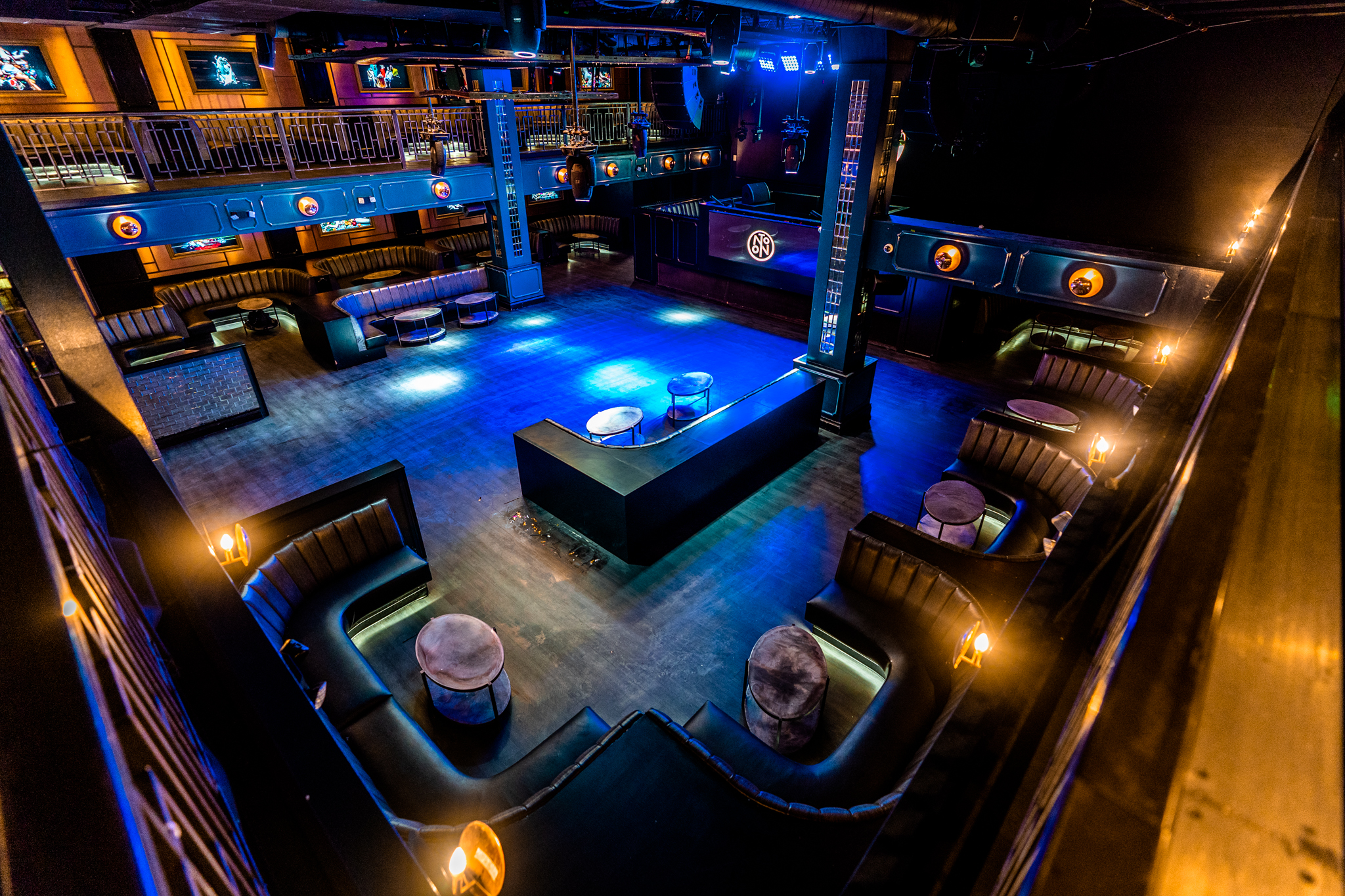 Interior of a modern nightclub with blue lighting, curved black leather booths, small round tables, and a central DJ booth with a NO ON sign.