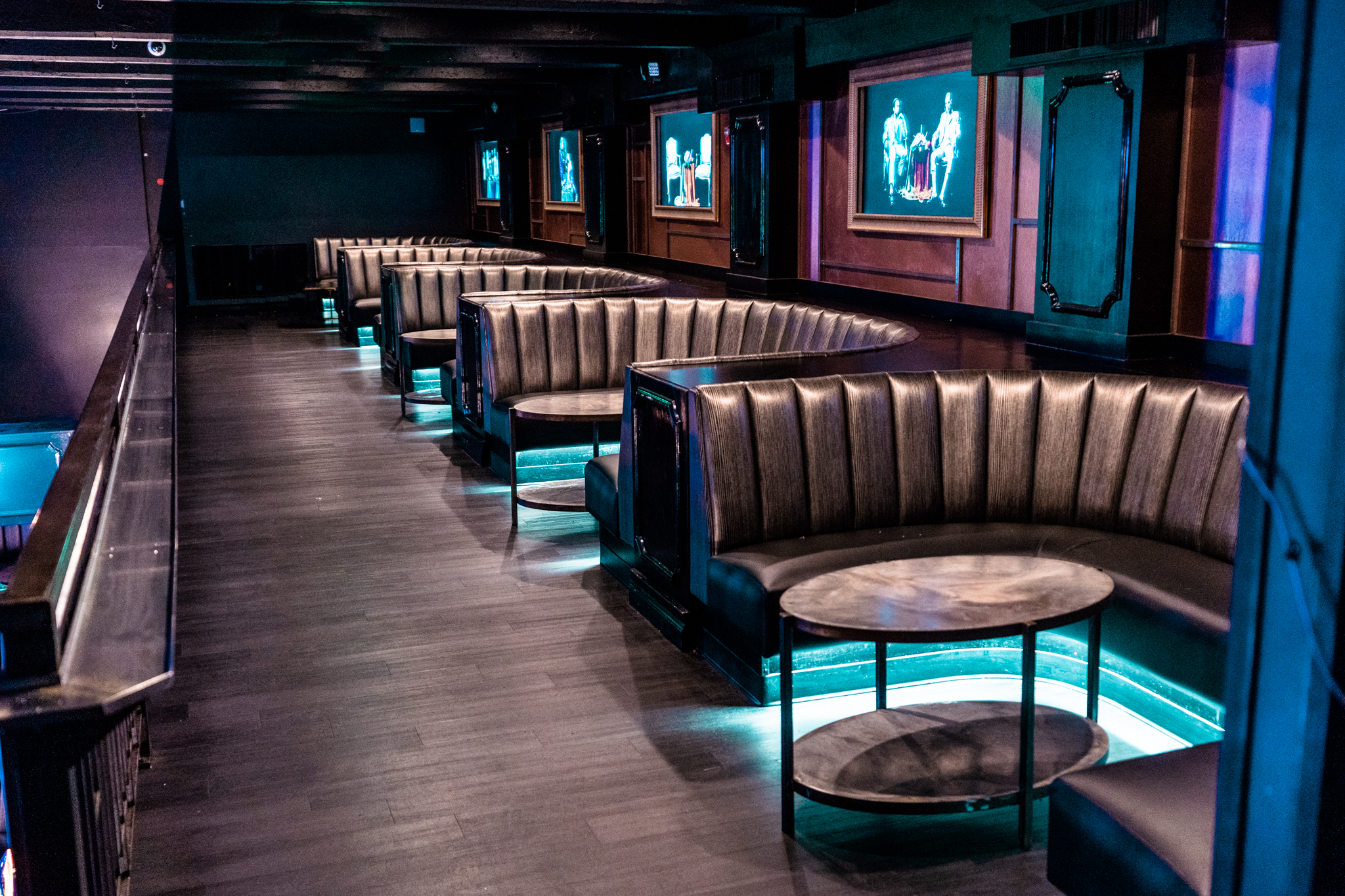 Row of curved gray leather booths with round wooden tables in a dimly lit lounge with blue accent lighting and framed artwork on the walls.