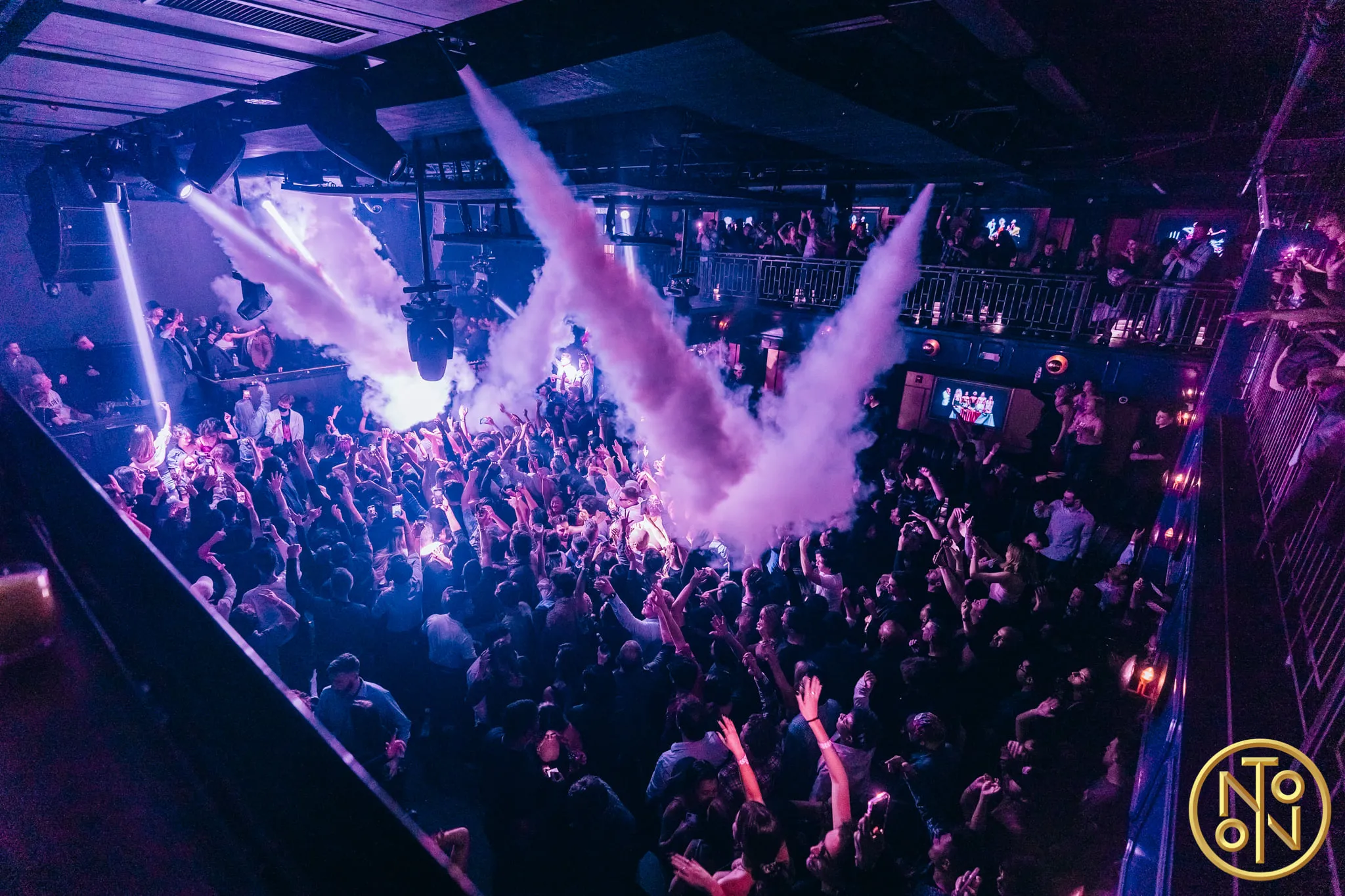 Crowded nightclub with people dancing and raising hands amid colorful lights and smoke effects.