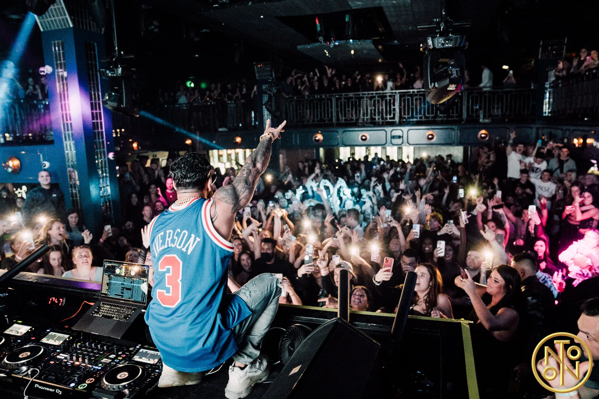 DJ wearing a blue Iverson basketball jersey with number 3 performing on stage, facing a large, excited crowd holding up phones with flashlights on.