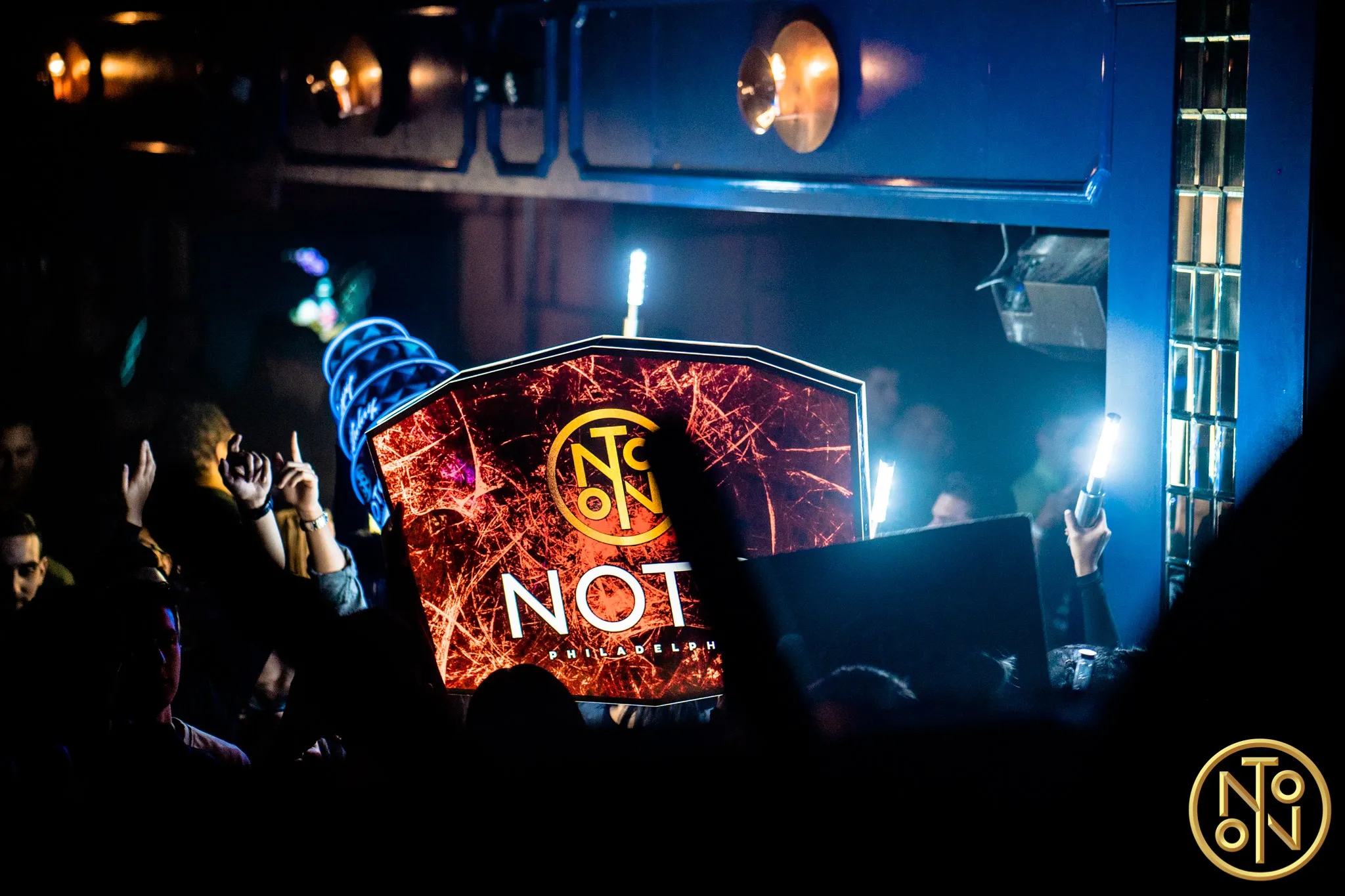 Crowd holding illuminated signs and light sticks in a dark club environment with a visible 'NOTO Philadelphia' sign.