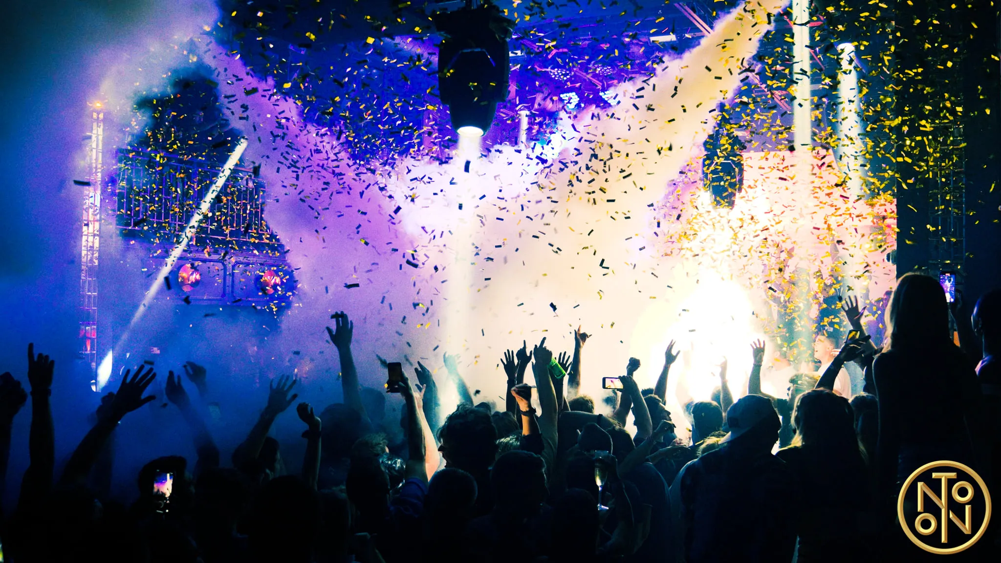 Crowd at a concert with raised hands, bright stage lights, smoke, and confetti in the air.
