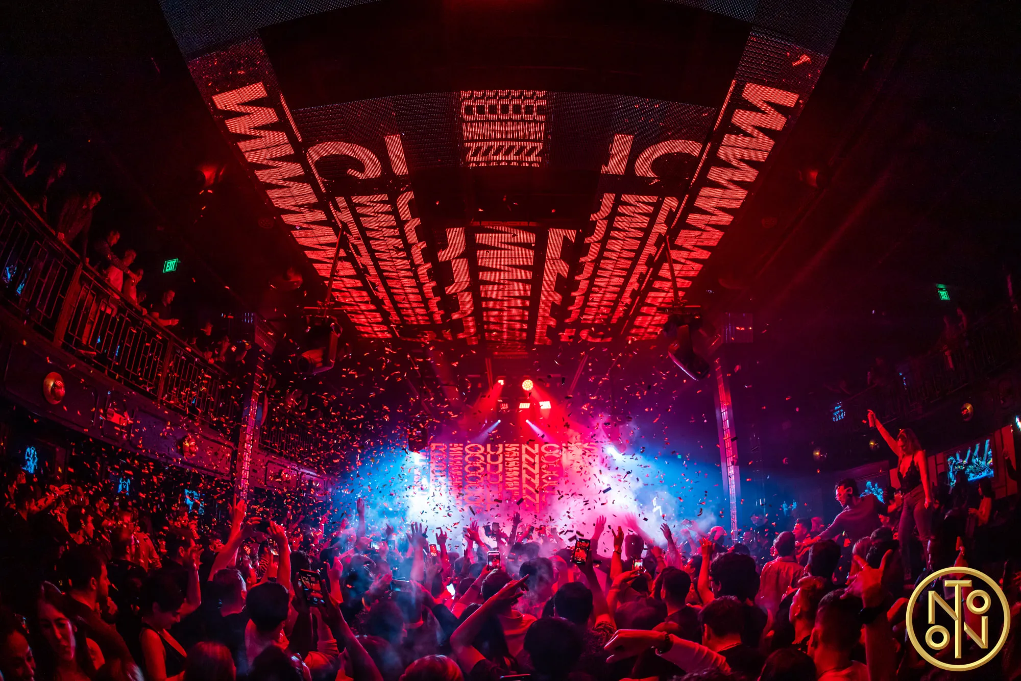 Crowded nightclub with red and blue lights, confetti falling, and people dancing and raising hands.