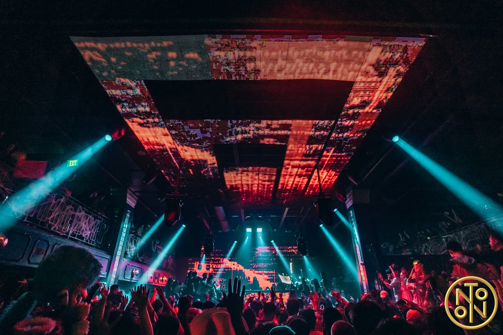 Crowded nightclub with colorful light beams and digital screens on the ceiling and stage.