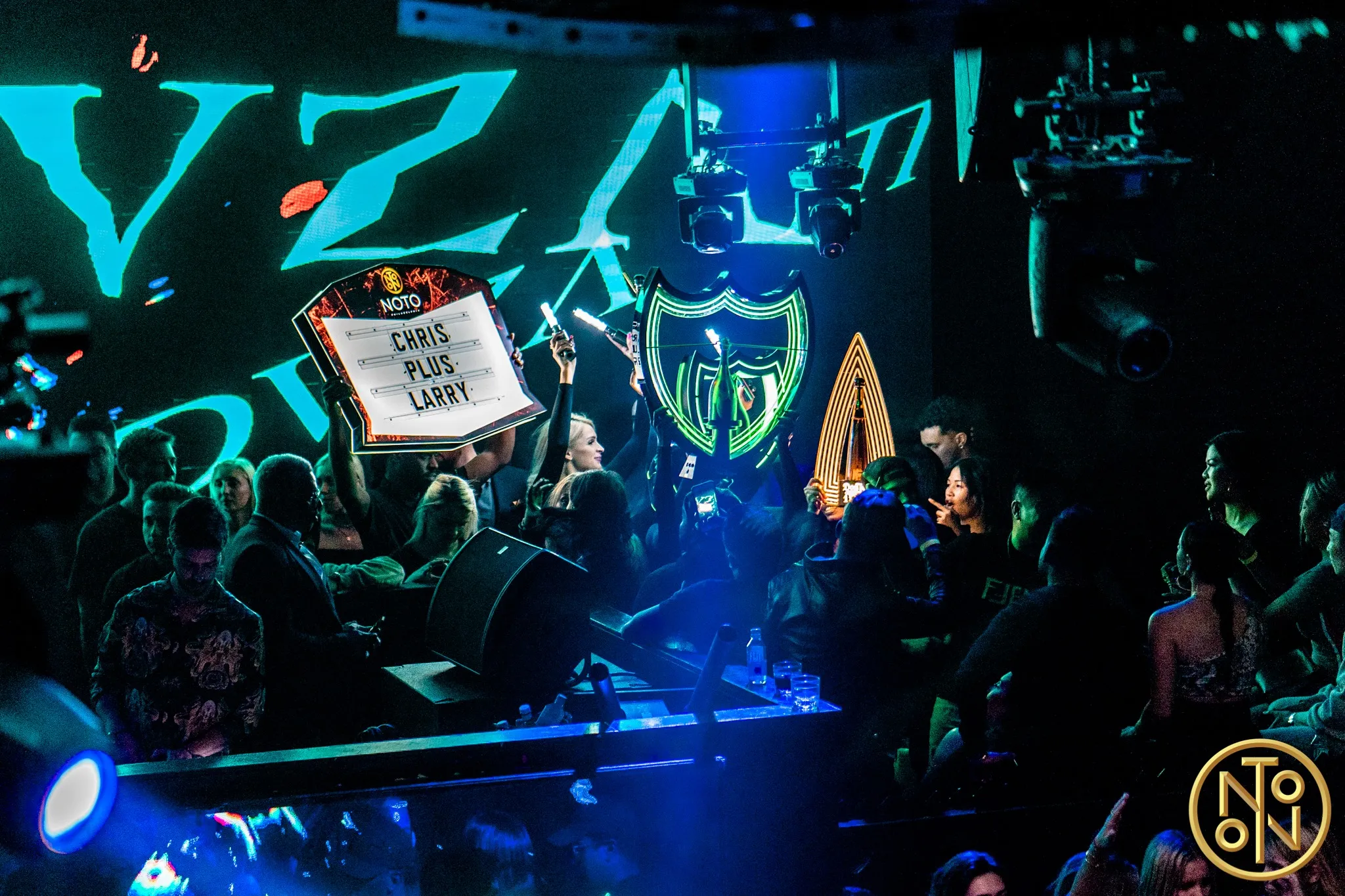 Crowd at a club with illuminated signs showing 'Chris Plus Larry' and neon logos, vibrant blue and green lighting.